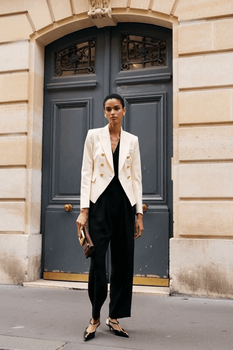 AI styling visual of an adult woman in a pale fitted jacket and black trousers, wearing black chain slingbacks and carrying a deep-red clutch.