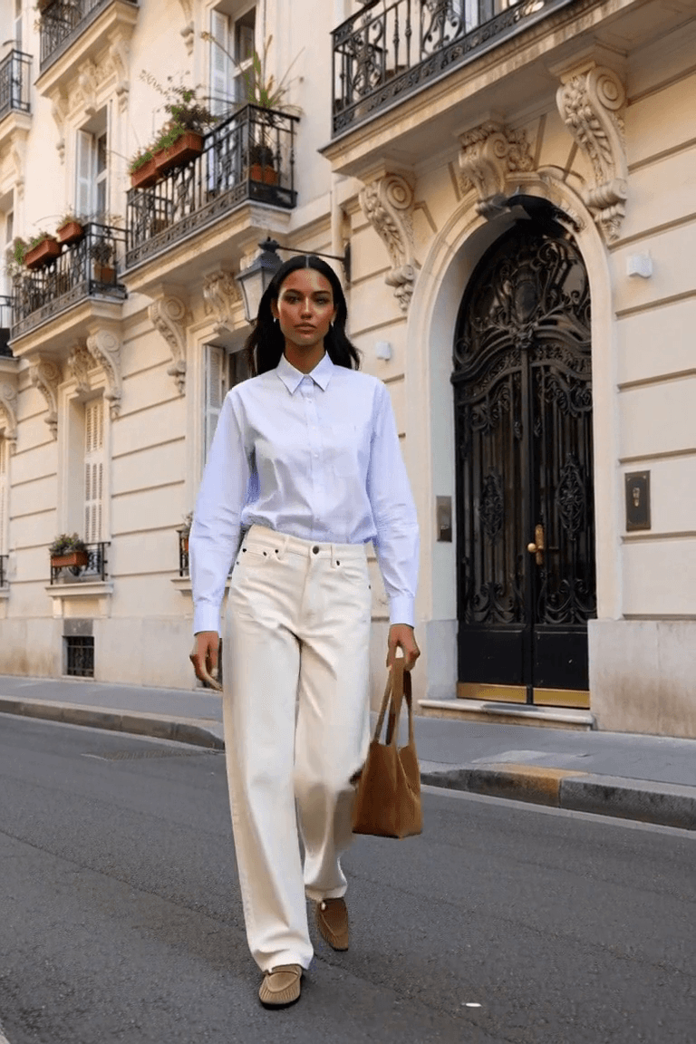 AI styling film of an adult woman in a pale striped shirt, white jeans, tan loafers and a brown bag on a stone-fronted city street.