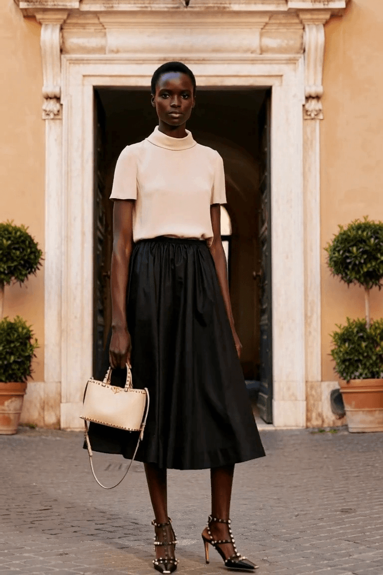 AI styling film of an adult woman in a blush mock-neck top, full black midi skirt and studded black pumps carrying an ivory handbag.