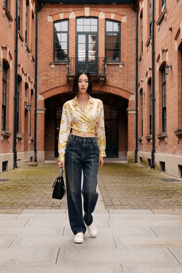 AI styling film of an adult woman in a gold-and-ivory printed wrap shirt, dark jeans, white trainers and a black bag in a brick courtyard.