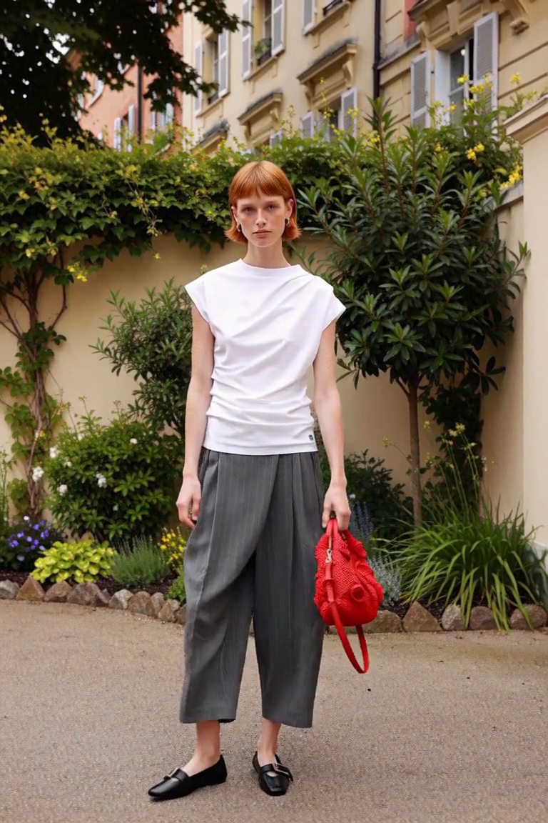 AI styling film of an adult woman in a white draped top, grey pinstripe trousers, black buckle flats and a red crochet bag beside a city garden.
